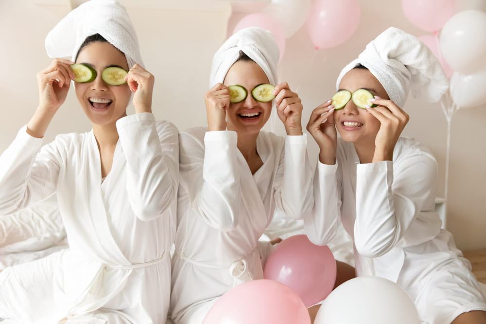 Friends in white robes enjoying a spa night at home with cucumber eye masks
