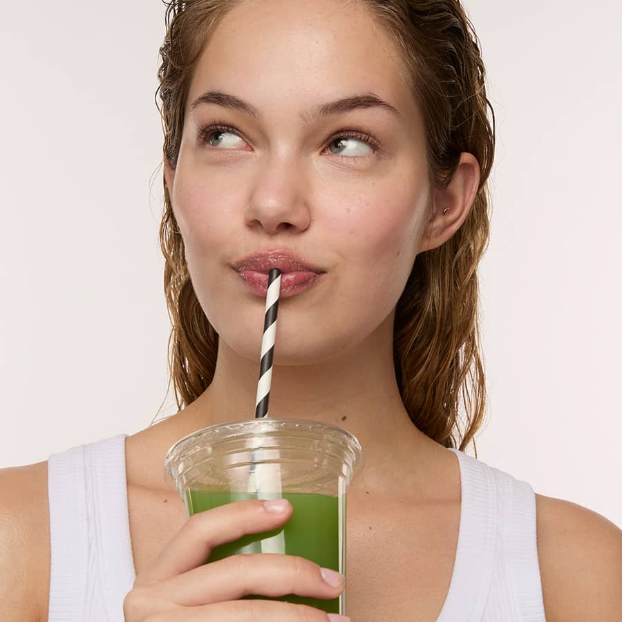 A woman with wet hair looks up and to the side while drinking from a clear cup with green liquid and a black-and-white striped straw against a plain light background.