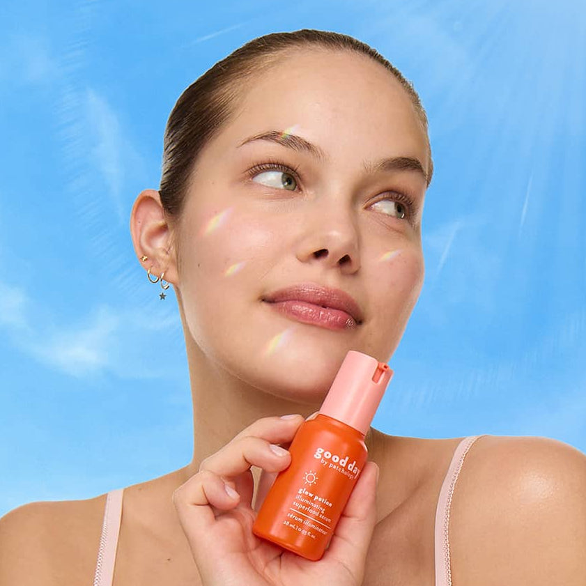 A woman with dabs of cream on her face holds an orange bottle of glow potion against a bright blue sky background.