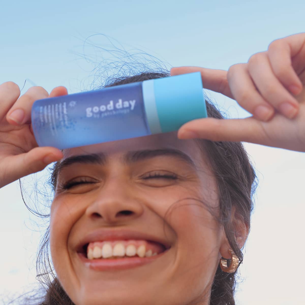 A smiling woman outdoors holds a blue and light blue crystal clear bottle over her forehead. The bottle has the text good day visible on the side.