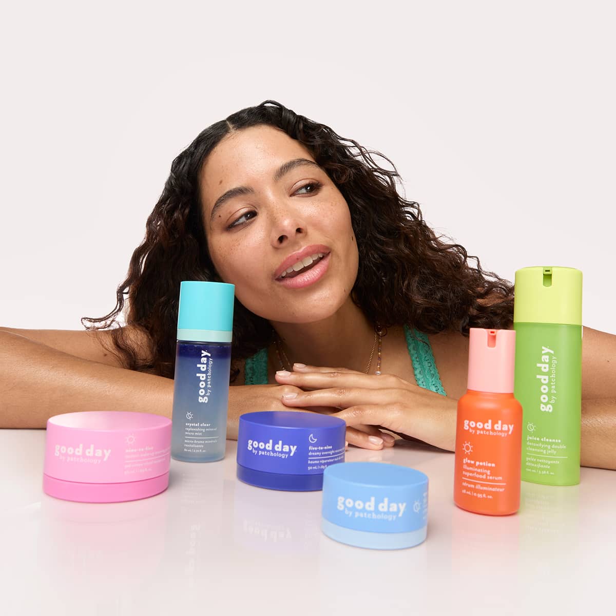 A woman with curly dark hair leans forward behind a group of Good Day skincare products on a white surface against a light gray background. Visible products include a pink nine-to-five jar, a blue five-to-nine bottle, a blue jar, a light blue eye pillow jar, an orange glow potion bottle, and a green juice cleanse bottle.