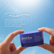 Hands holding a blue cosmetic five-to-nine jar labeled 'good day' against a bright blue sunburst background with white text describing molecular sunshine benefits.