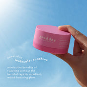 A hand holding a pink round cosmetic jar labeled 'good day' against a bright blue sky background, with marketing text on the left.