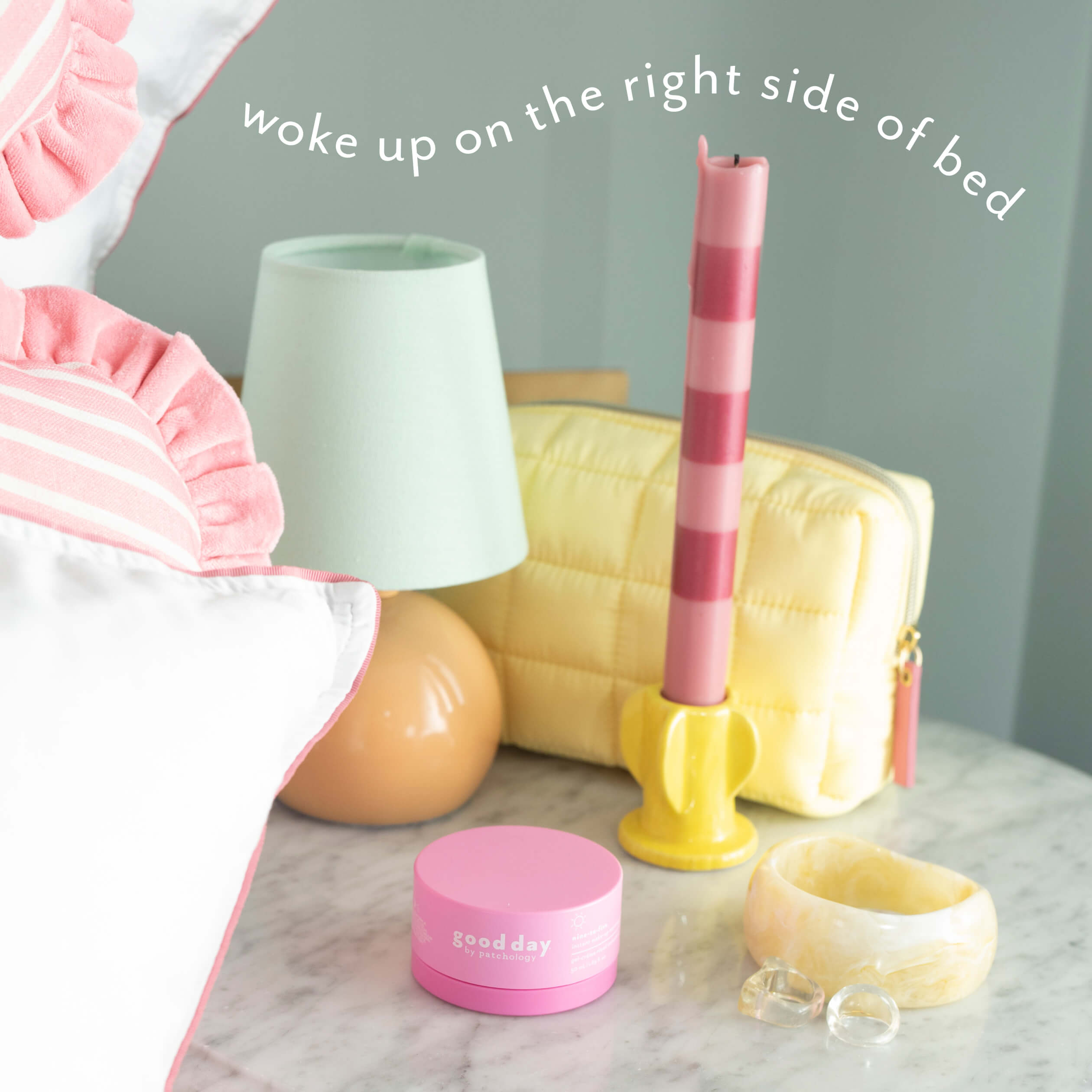 A styled bedroom scene with pink and white bedding, a pale green lamp, a yellow quilted pouch, a pink striped candle in a yellow holder, a pink jar labeled Good Day, a translucent cream bowl, and clear rings on a marble surface. White script text at the top reads 'woke up on the right side of bed'.