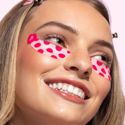 A close-up of a smiling woman with pink and white polka-dot eye patches applied under her eyes against a light pink background.