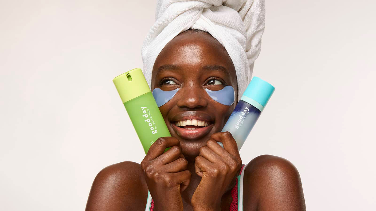 A smiling woman with a white towel wrapped around her head holds two cleanser and mist skincare bottles, one green and one light blue, against a plain light background.