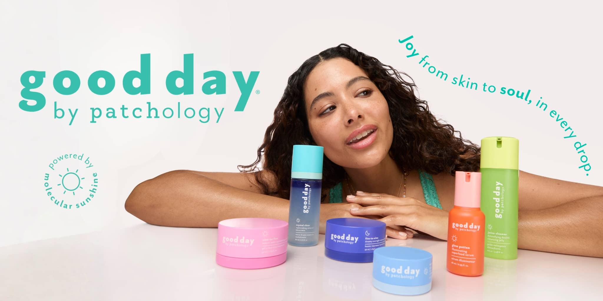 A woman with curly hair rests her head on her arms on a white surface, surrounded by several Good Day by Patchology skincare products in various colors including pink, blue, orange, and green. The background is a clean white with teal brand text and a small circular logo.