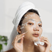 Woman applying a face mask with a white towel on her head against a neutral background