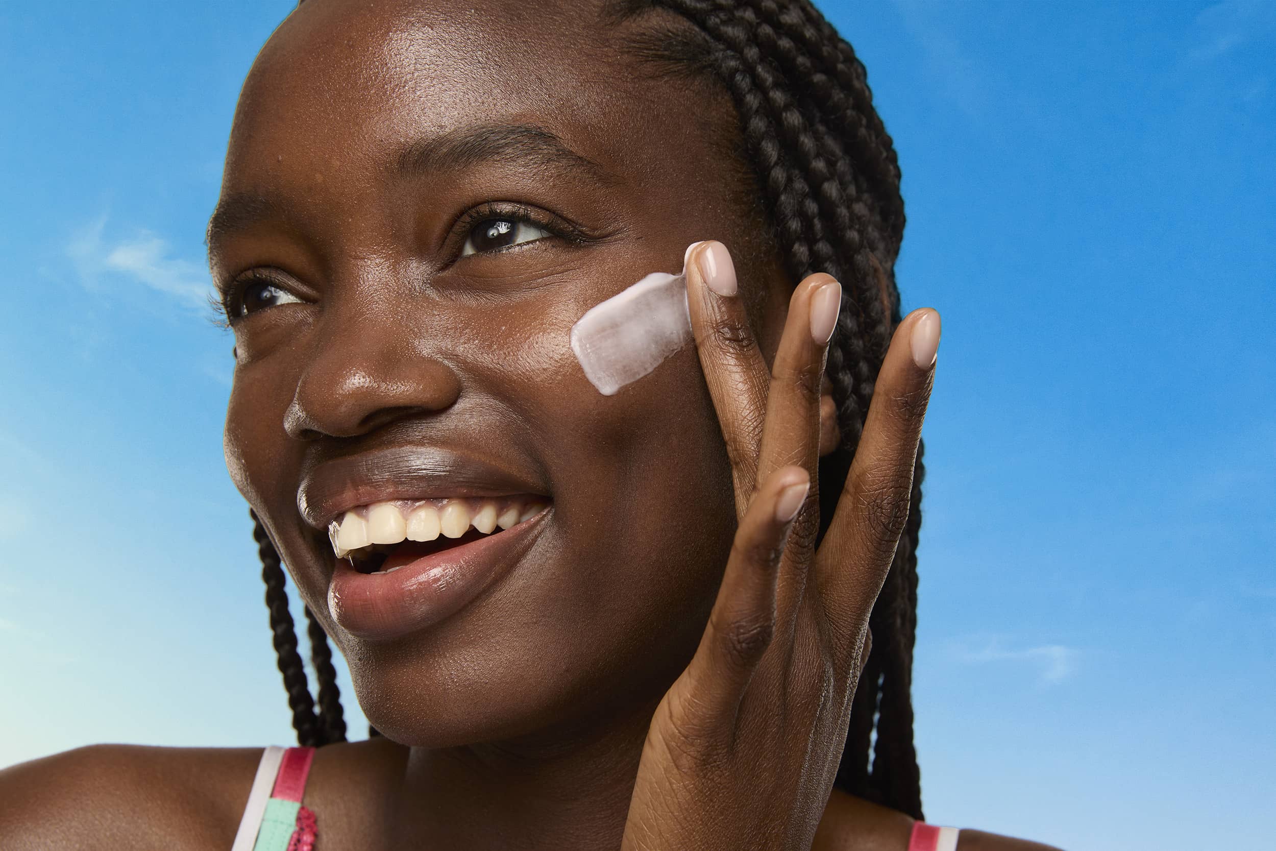 A smiling woman with braided hair applies white nine-to-five moisturizer cream to her cheek against a bright blue sky background.