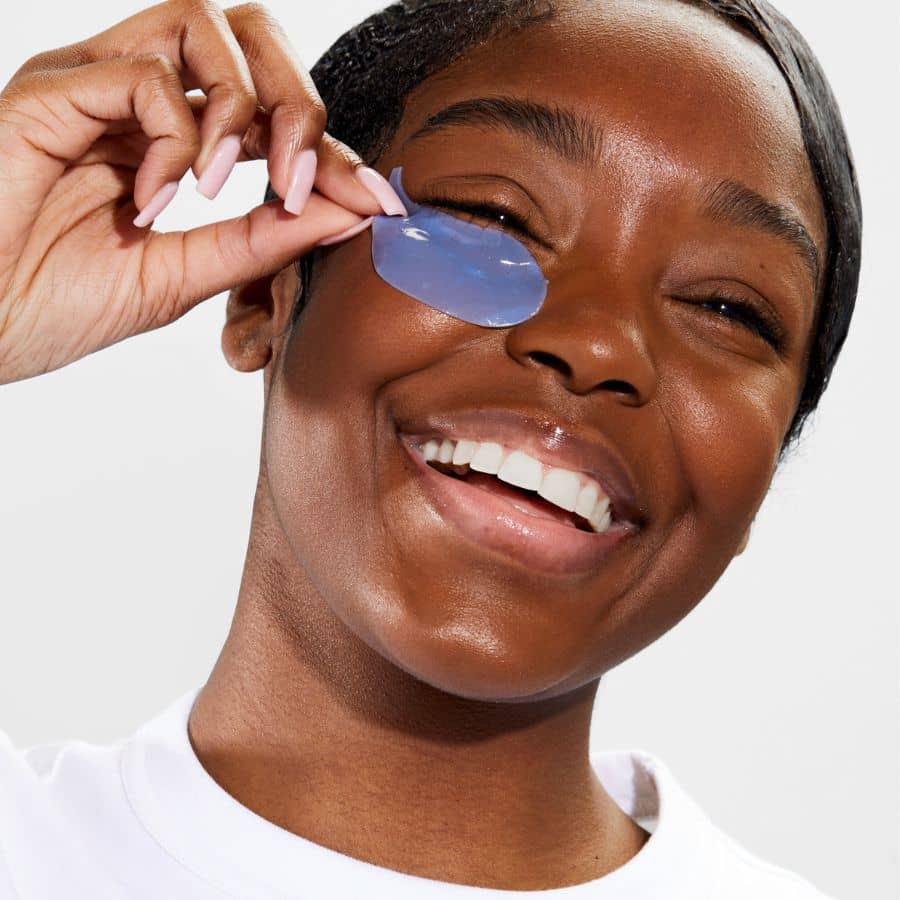 Woman placing blue hydrogel under-eye patch, smiling during brightening eye care routine
