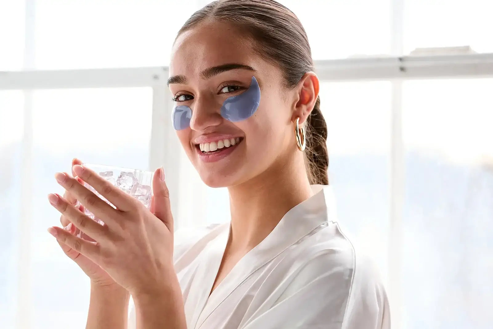Smiling woman wearing blue under-eye gel patches and holding ice, cooling skincare routine for brightening eyes

