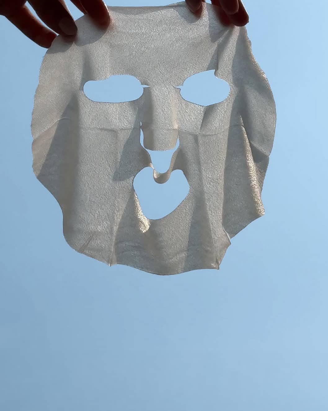 hands holding sheet mask against a bright blue summer sky