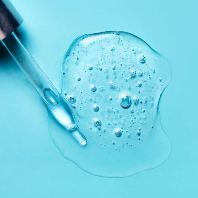 Clear facial serum with tiny bubbles and a glass dropper on a vibrant blue background for skincare application.