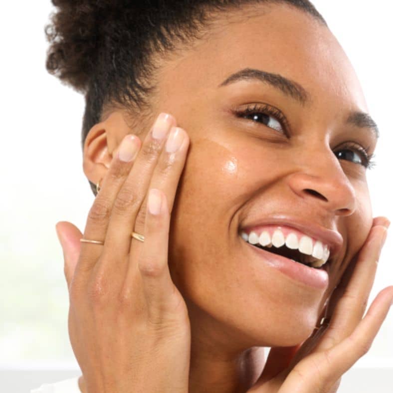 Smiling woman applying a hydrating skincare serum to her cheek for a radiant and smooth complexion.
