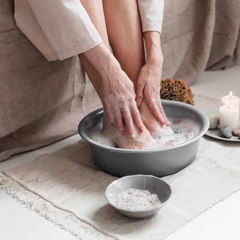 Person soaking and gently scrubbing feet in a warm, foamy foot bath with candles and spa essentials nearby
