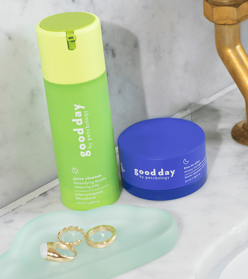 Two Good Day skincare products on a marble bathroom counter: a bright green juice cleanser bottle and a royal blue cream jar, with gold rings on a mint tray nearby.