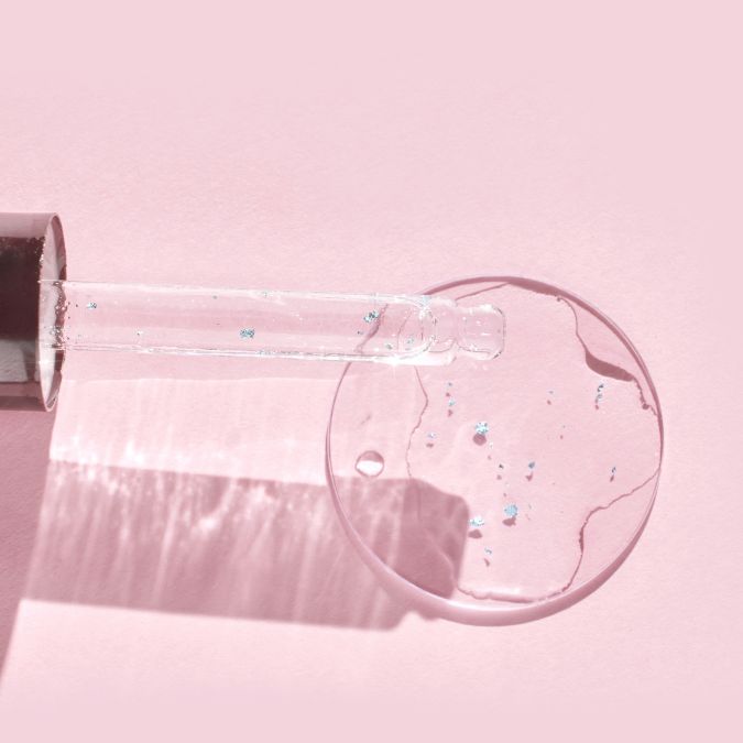 Glass dropper dispensing clear serum with shimmer particles onto pink surface for hydrating skincare application